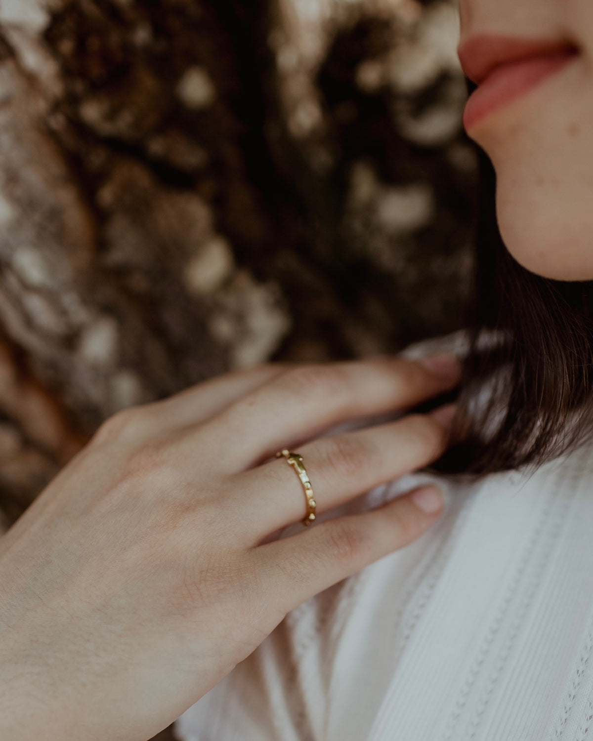 A close-up lifestyle shot of a woman wearing a delicate gold rosary ring on her finger, with her hand resting on her chest against a soft-focus background.