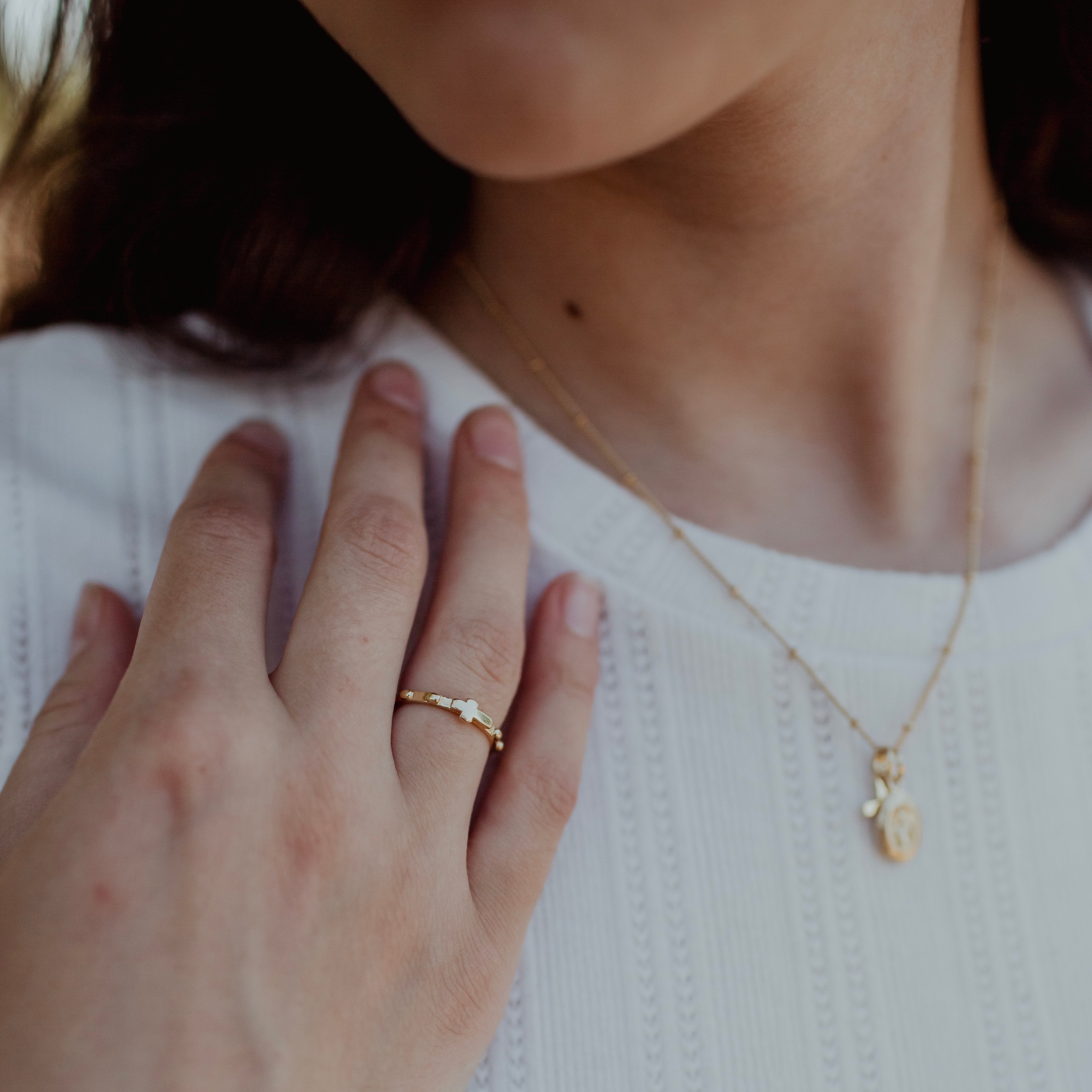 A close-up lifestyle shot of a young woman wearing a delicate gold rosary ring on her finger, paired with a white ribbed knit top and a gold cross and miraculous medal necklace.